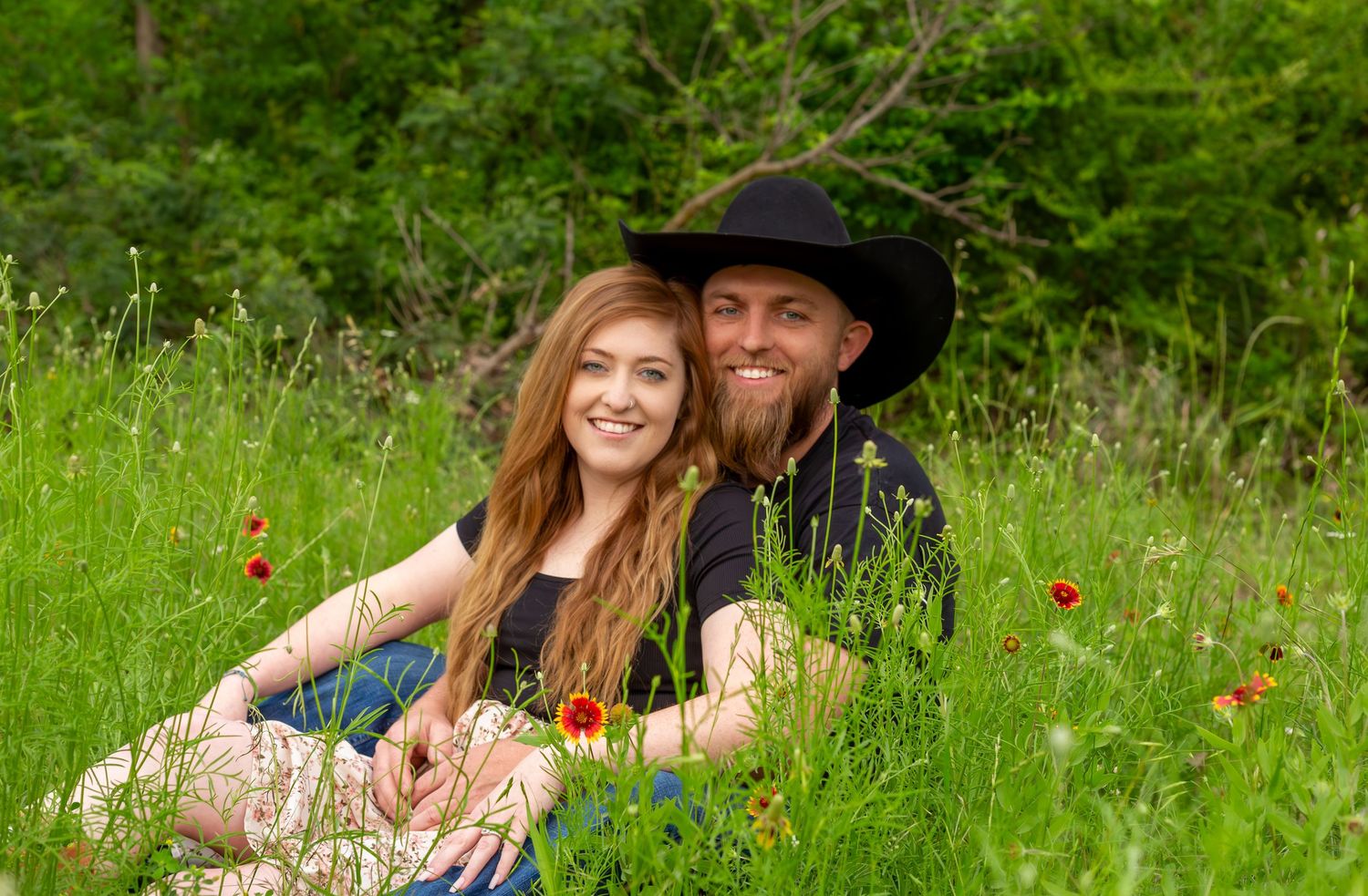A couple embraces in a lush green meadow filled with wildflowers during a sunny summer day.