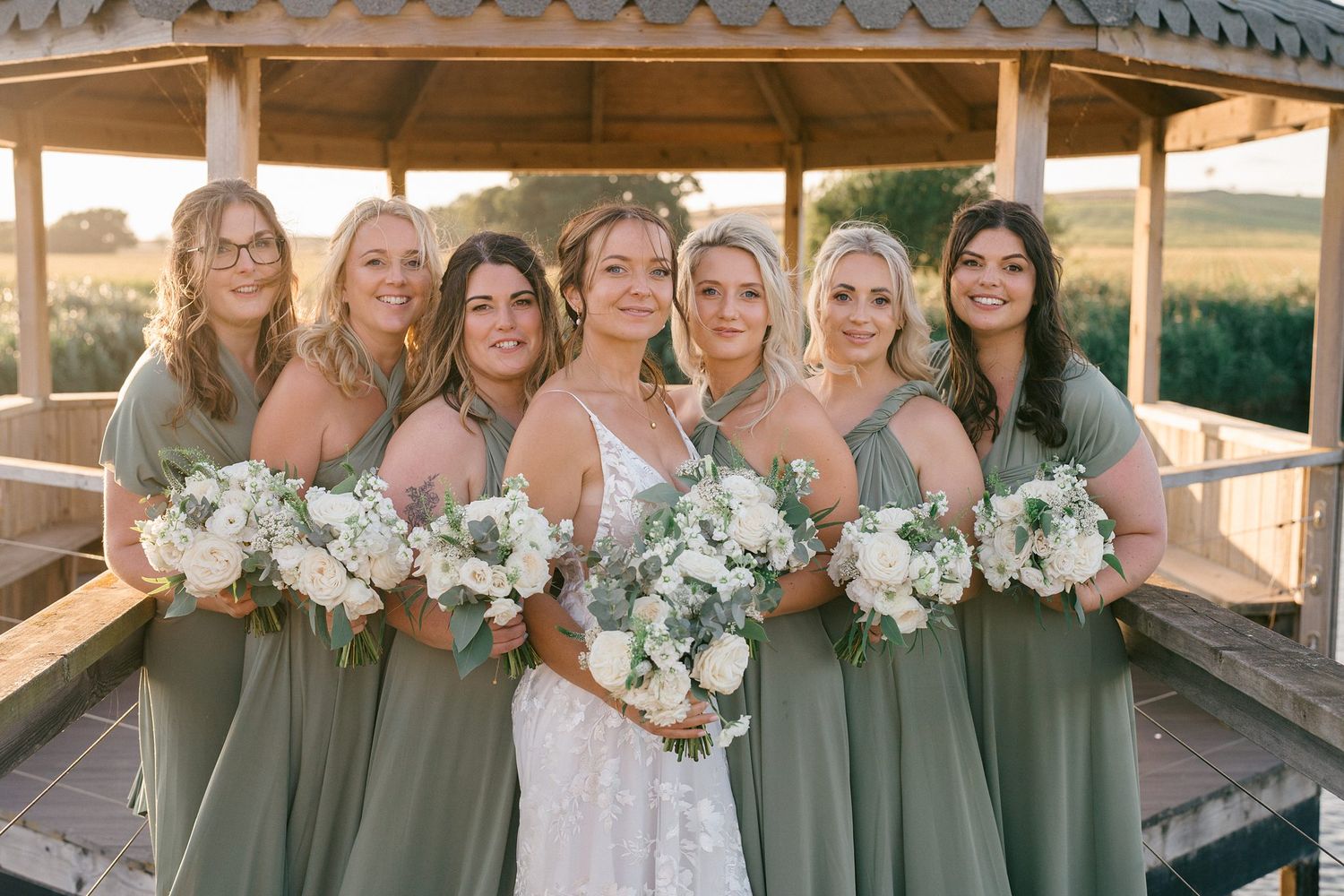 Wedding party in sage green dresses holding white floral bouquets pose together outdoors at sunset.