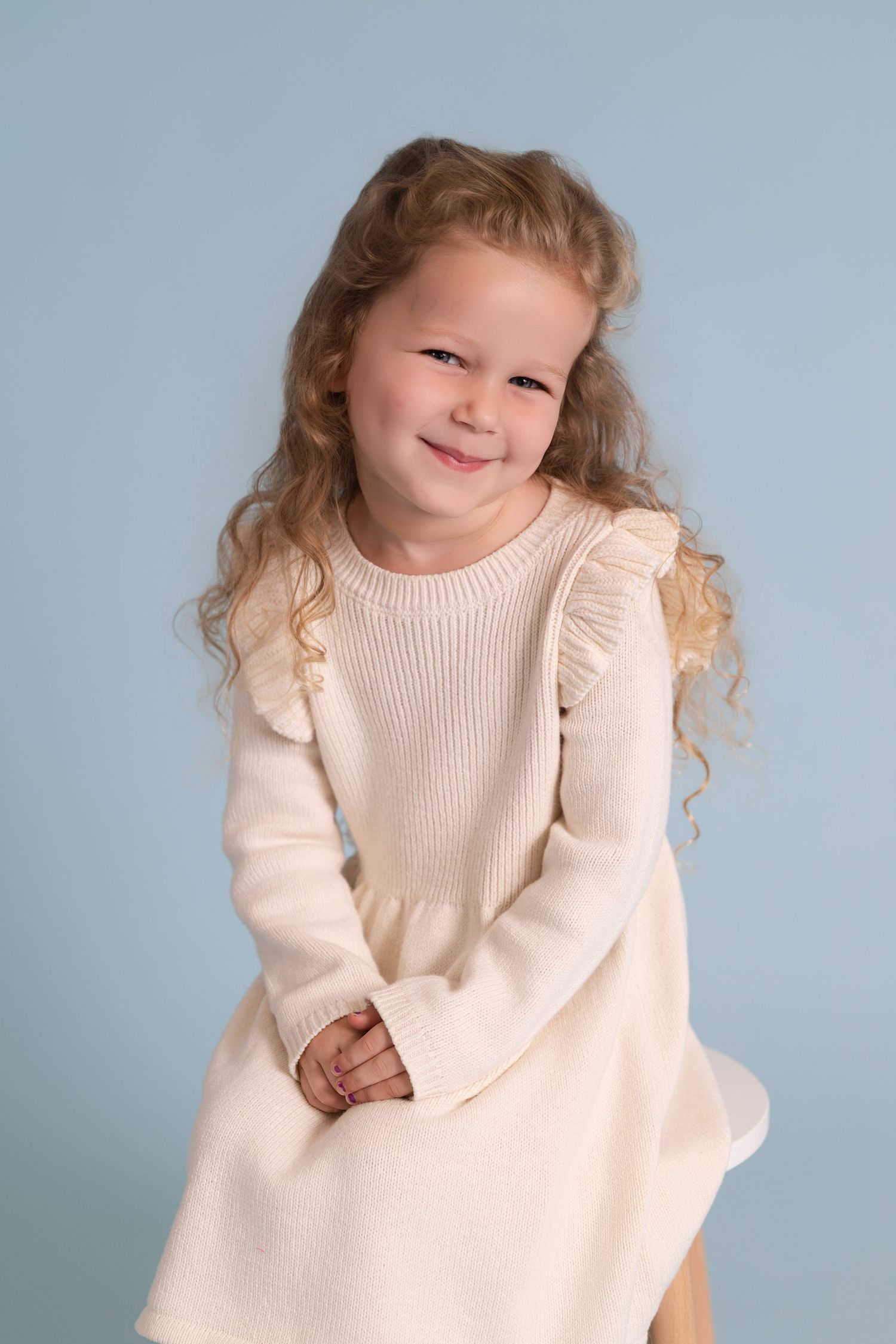 Child wearing cream colored knit sweater with ruffle details against blue background.