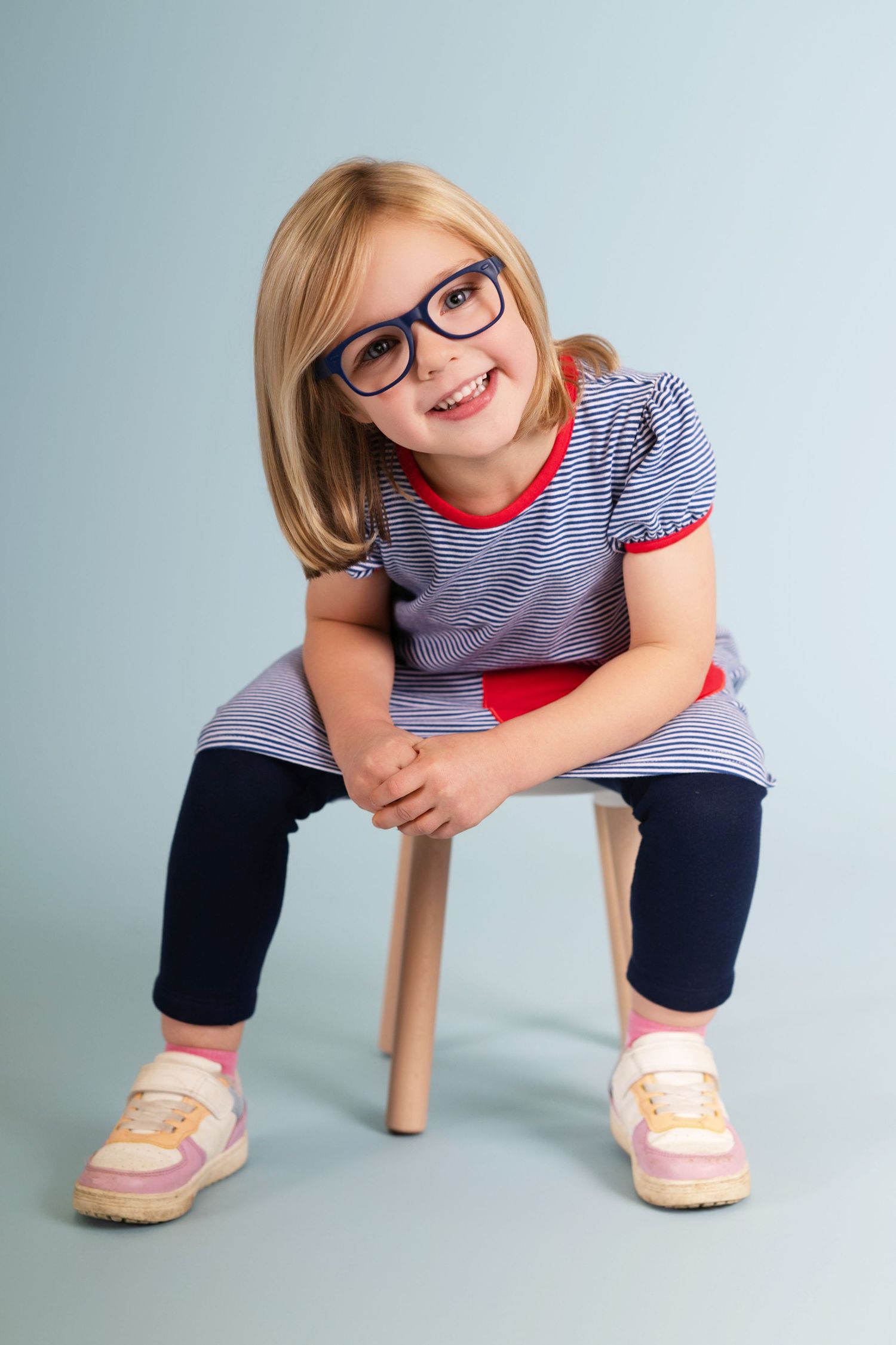 Child sitting on wooden stool wearing striped shirt and navy pants with pink shoes.