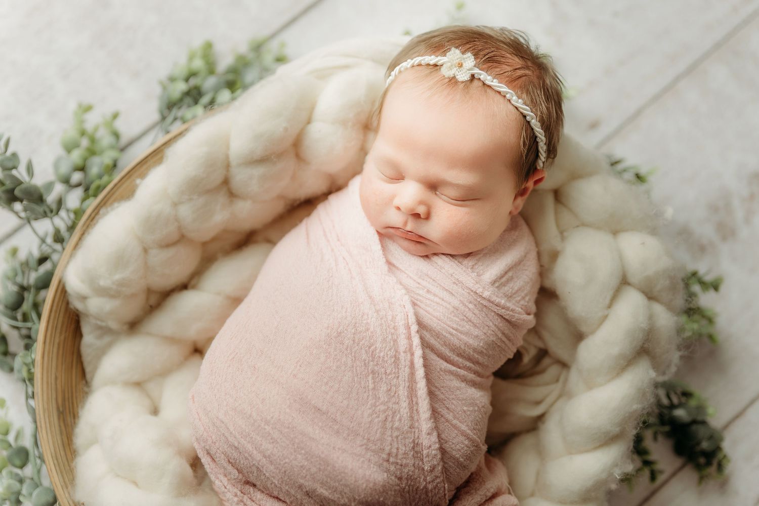 Newborn baby sleeping peacefully on white blanket wearing delicate pink wrap in professional portrait setting.