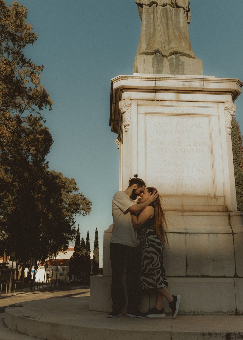 A couple embraces against a stone monument backdrop in warm afternoon sunlight.