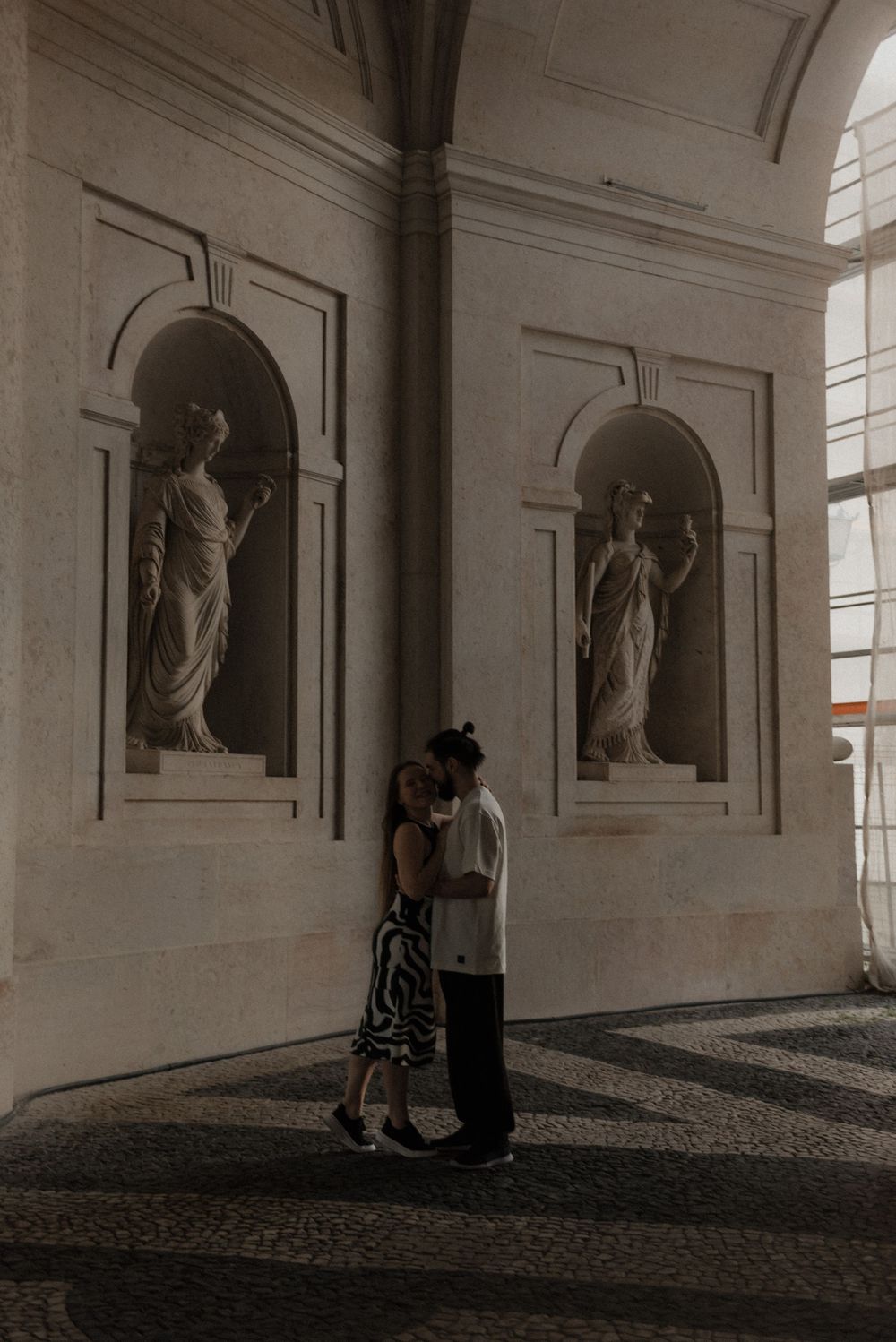 A couple shares an intimate moment beneath classical statues in an ornate architectural hallway.