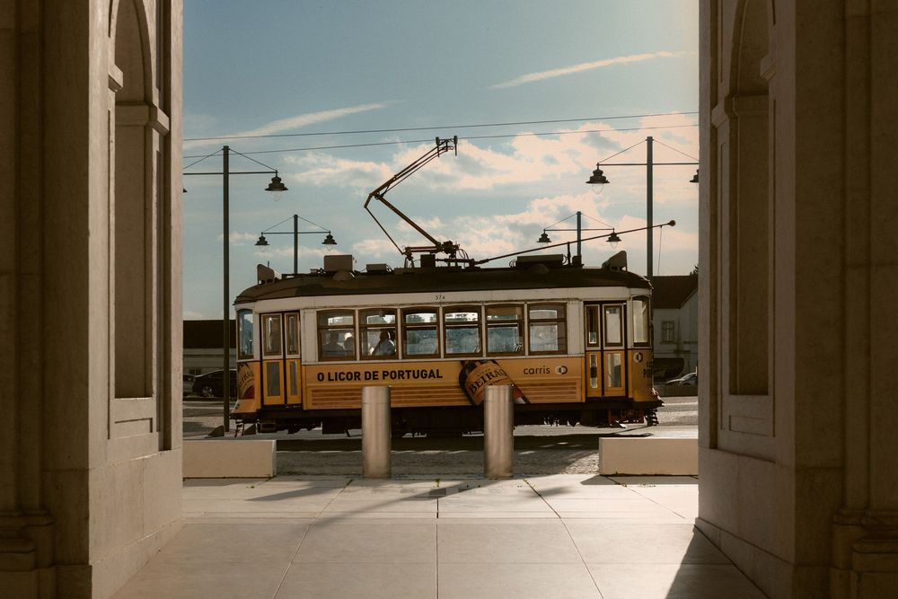 A vintage yellow tram car framed by classical architecture columns against a blue sky.