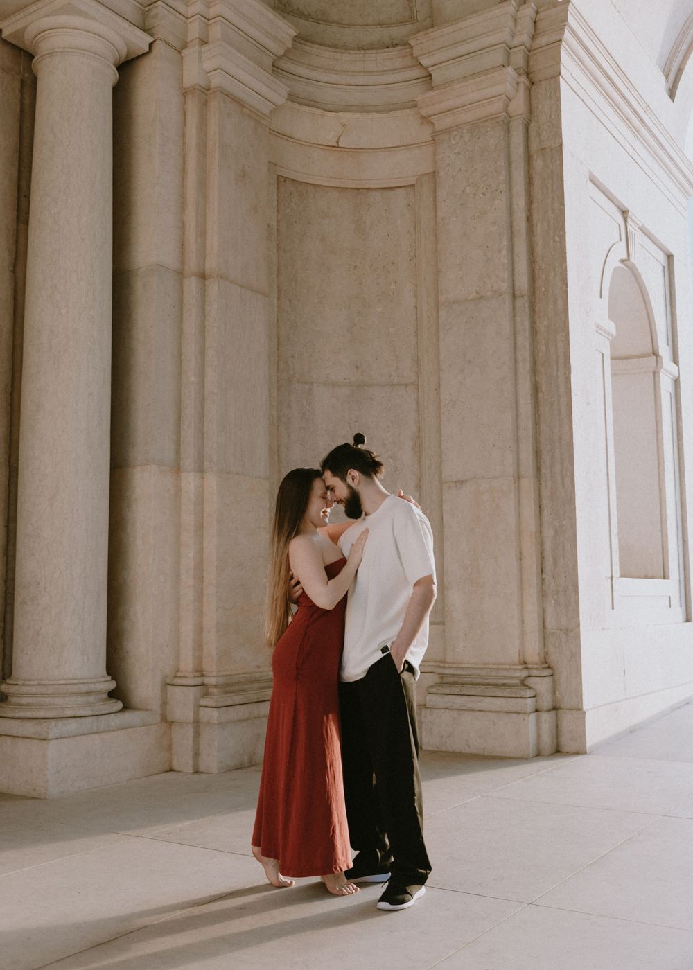 A couple shares a romantic moment between classical stone columns in formal attire.