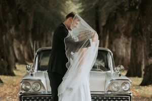 A romantic moment between newlyweds beside a classic vintage car in a tree-lined setting.