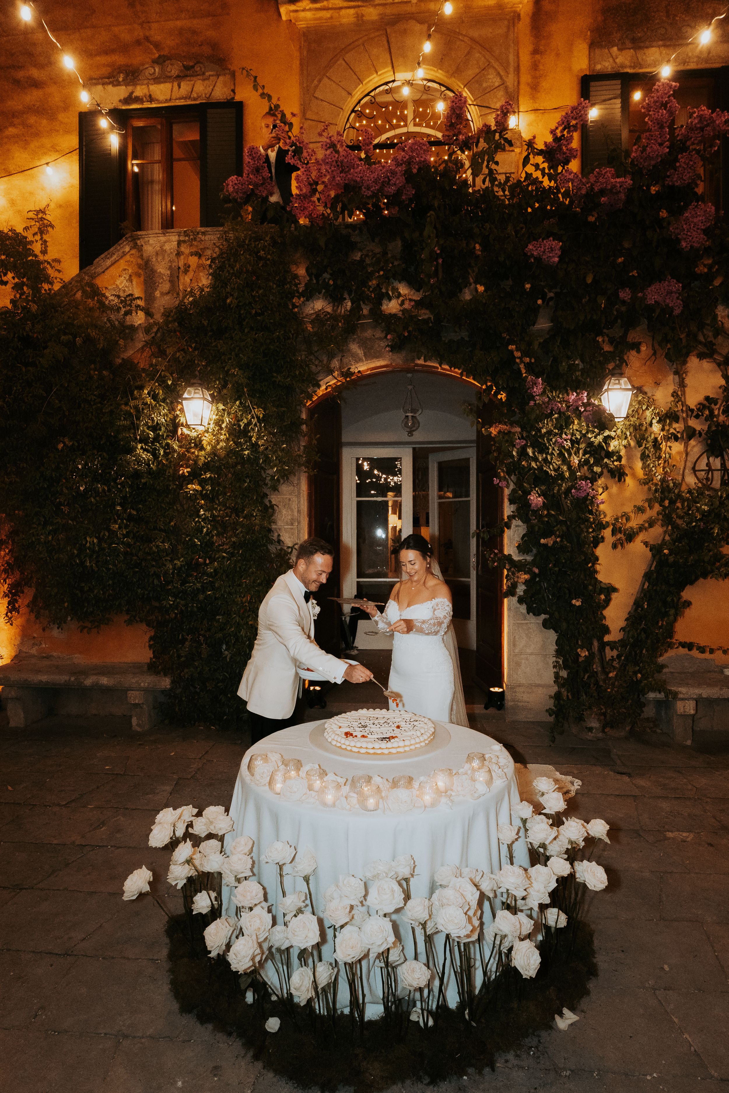 Elegant wedding cake display surrounded by white flowers and romantic lighting at reception.