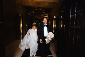 Newlyweds walk together in an elegantly lit hallway carrying a white floral bouquet.