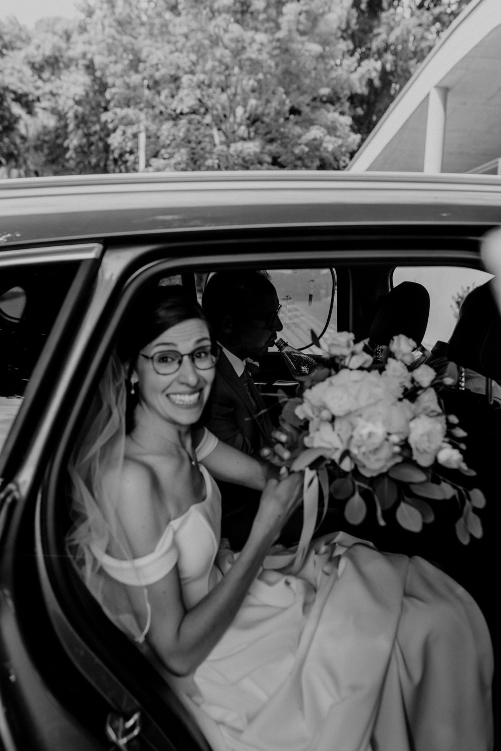 A black and white photo showing someone in a car with white flowers and a veil.