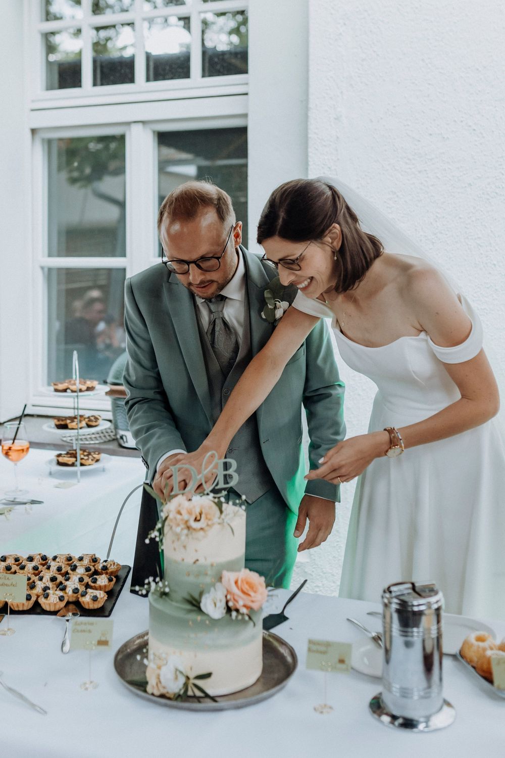 People in formal attire cutting a white wedding cake together at an elegant venue.