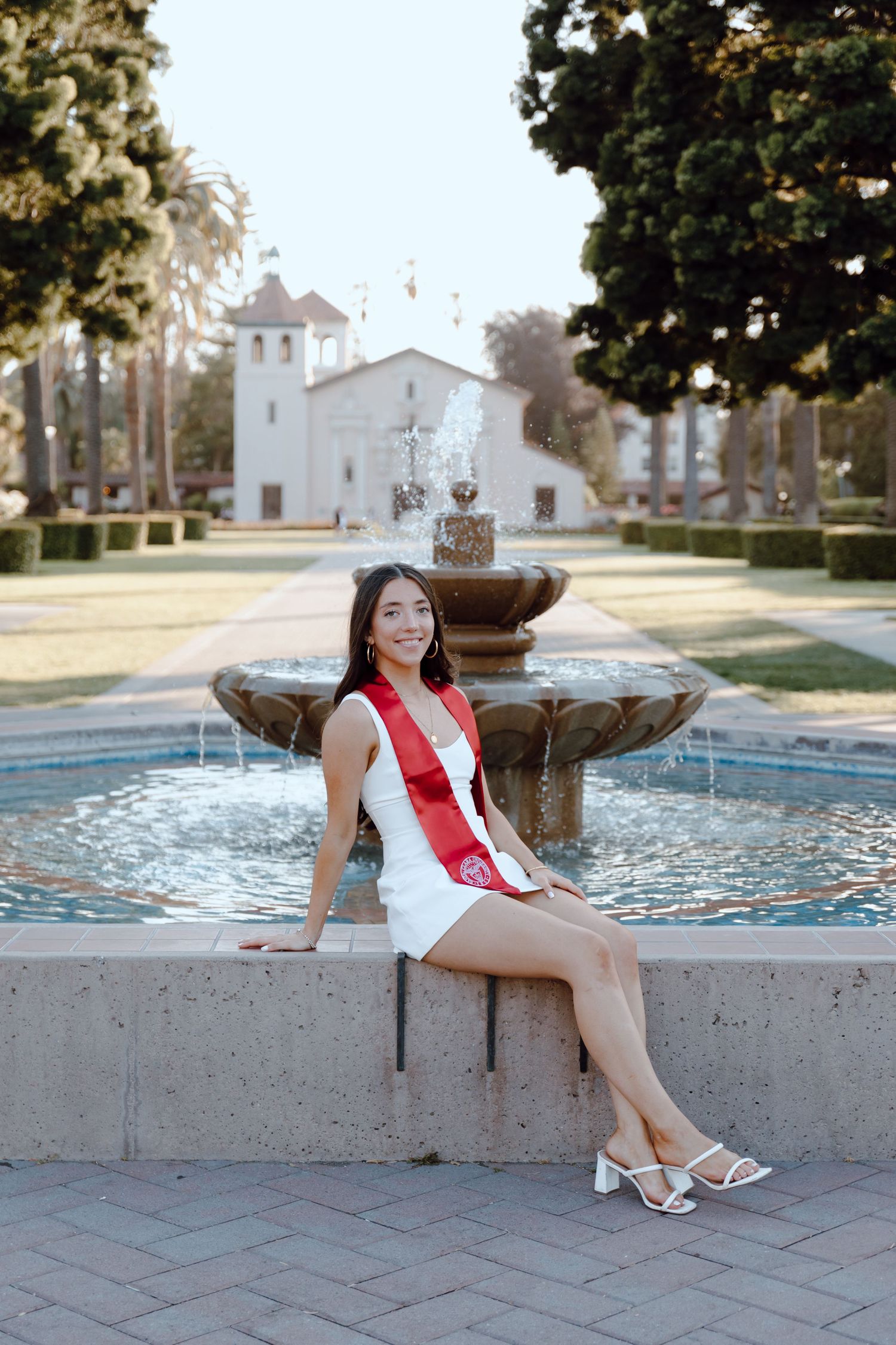 Santa Clara University graduate poses for her grad photoshoot by sitting on edge of fountain. Taken by Bay Area Graduation Photographer.