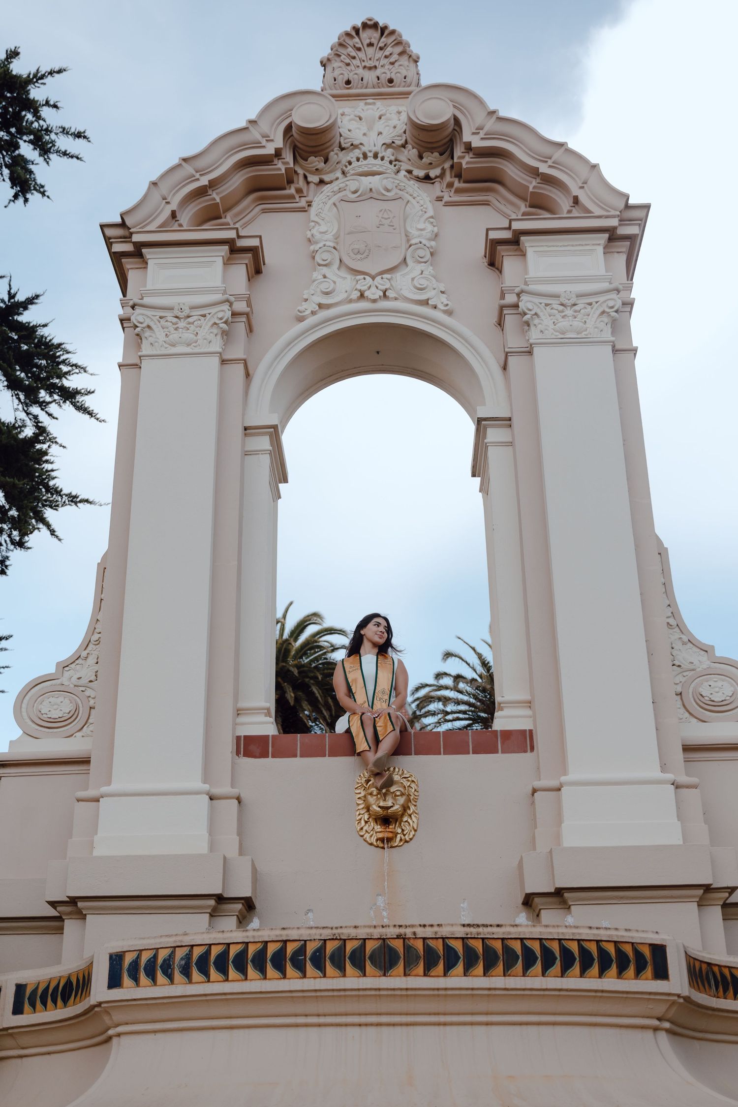 Graduate sits underneath an archway, looking off at the University of San Francisco. Grad photos taken by Bay Area Graduation Photographer.