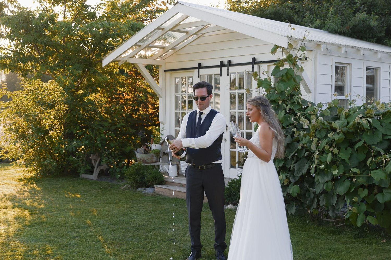 Groom pops champagne bottle as bride watches outside a charming white garden building.