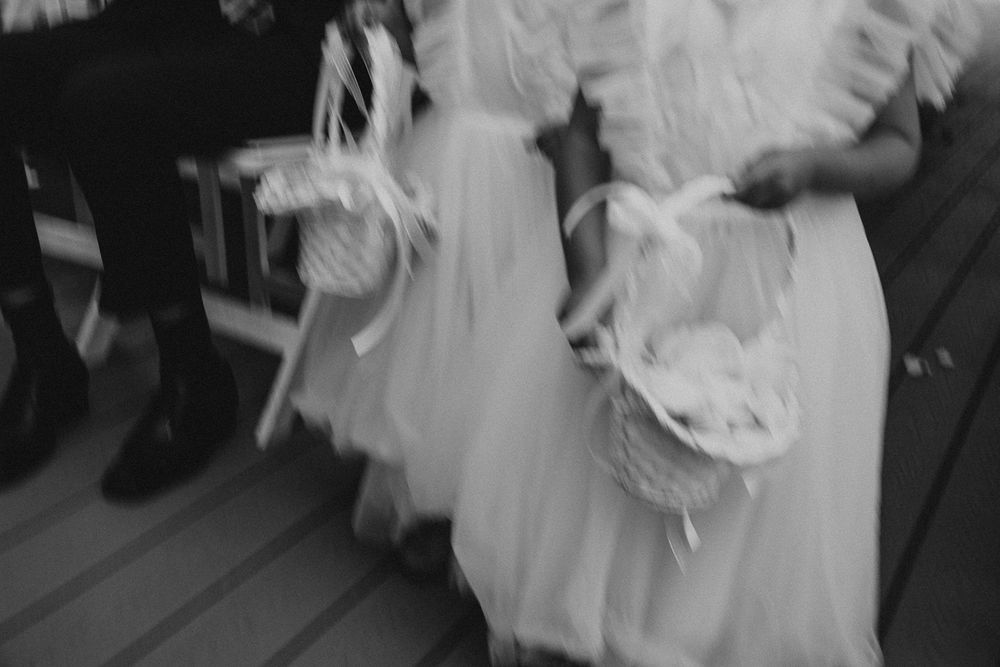 Blurry black and white photo of flower girls in white dresses carrying baskets at a wedding ceremony.
