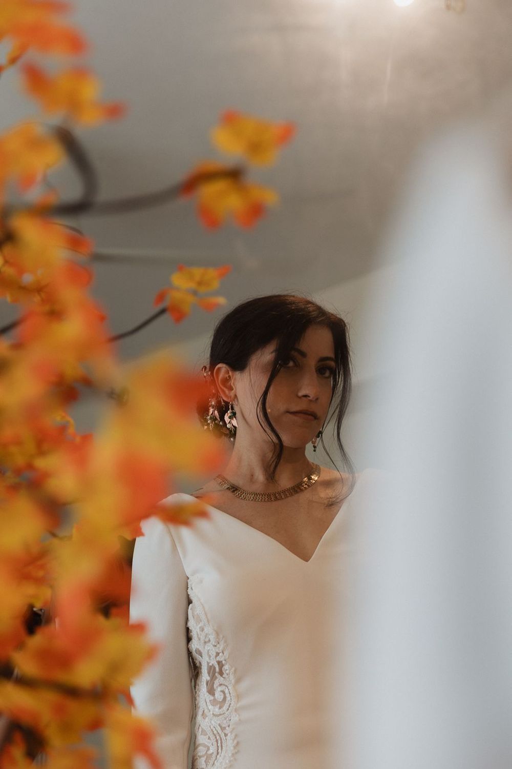 Bride in white lace wedding dress framed by vibrant orange and yellow autumn leaves indoors.