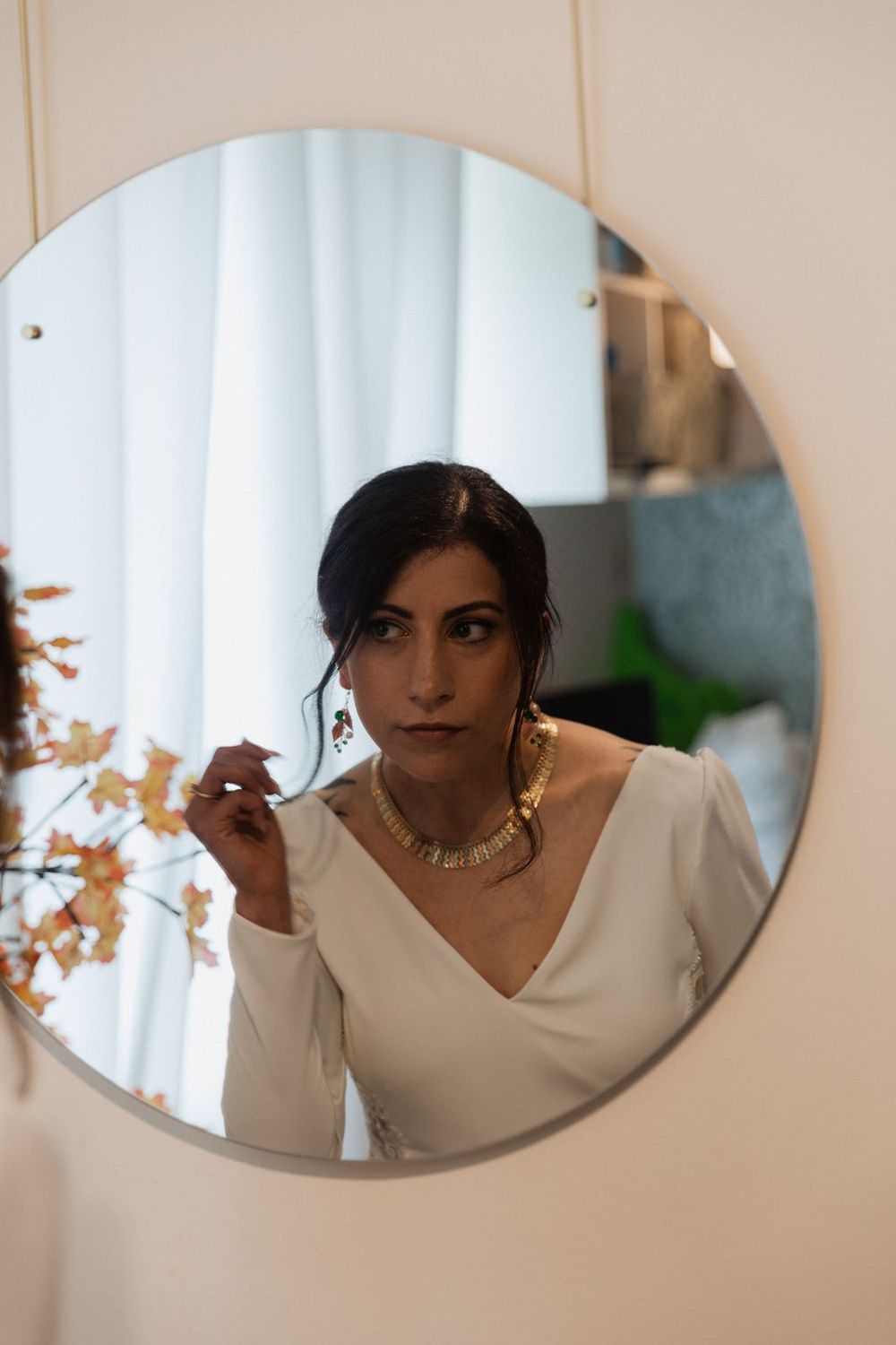 Bride in white V-neck wedding dress adjusting earring while reflected in a round mirror.