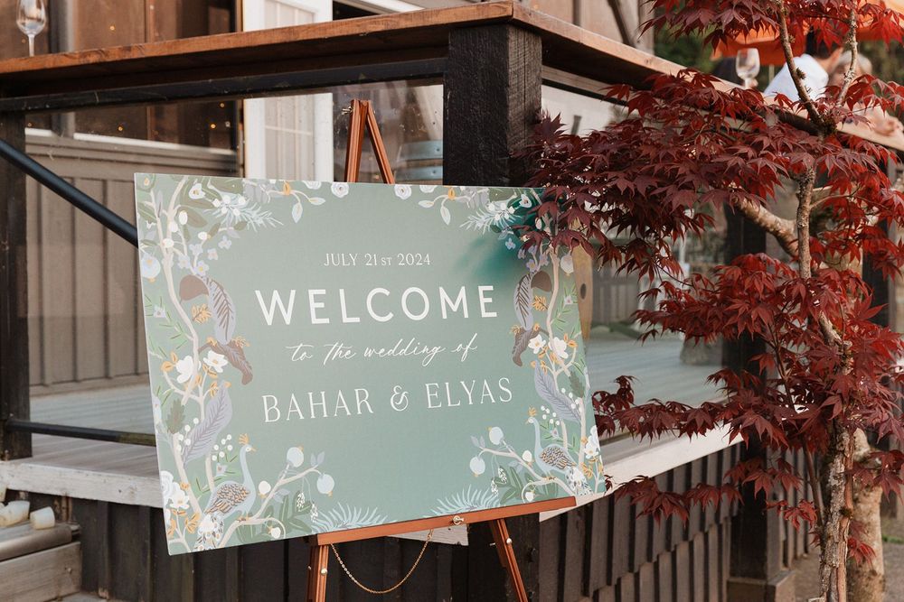 A elegant sage green welcome sign for the wedding of Bahar and Elyas displayed on a wooden easel.