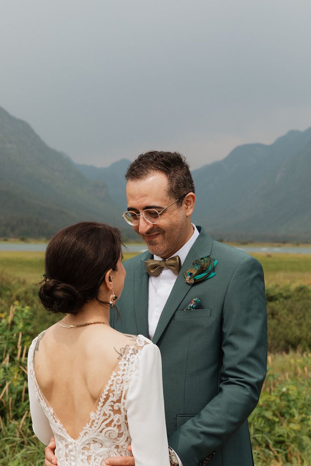 A groom in a teal suit with a peacock feather boutonniere smiles at his bride against a mountain backdrop.