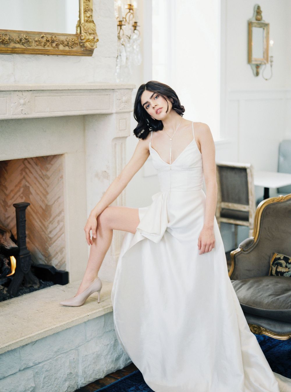 A bride in a white wedding dress sits elegantly on a blue velvet couch in an ornate room with a fireplace.