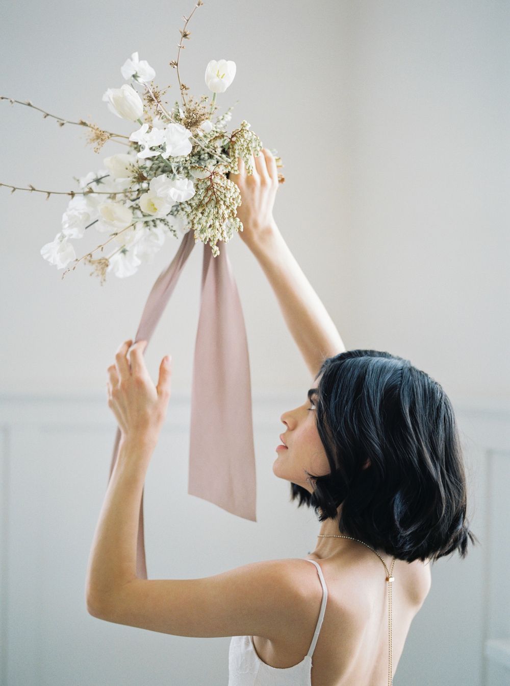 A person in a white dress reaches up gracefully to touch delicate white flowers and branches against a light background.