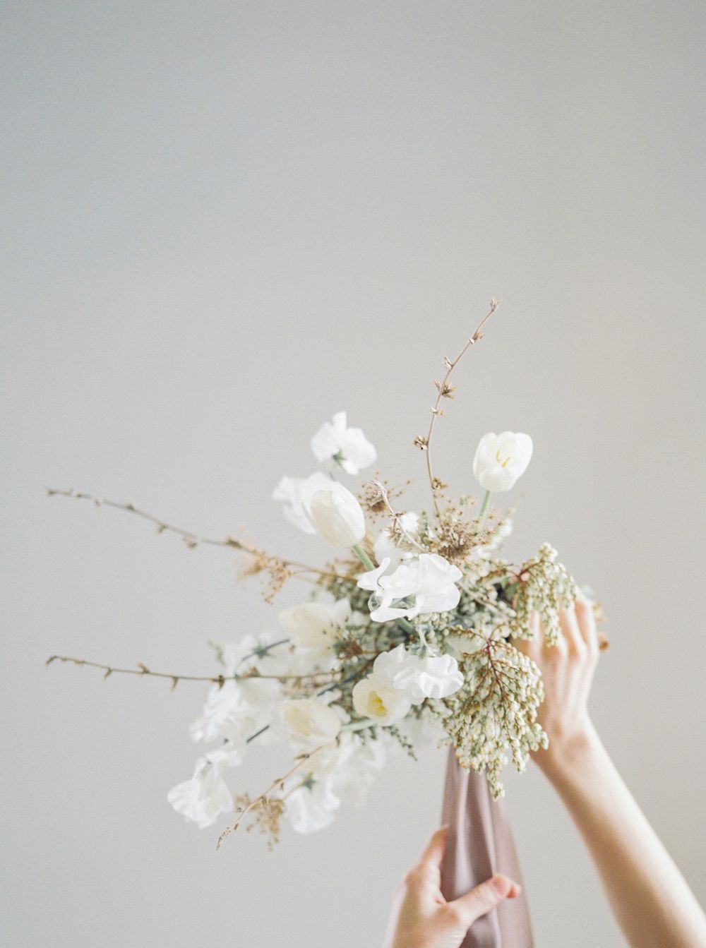 Hands holding a delicate white floral arrangement with dried branches against a light gray background.