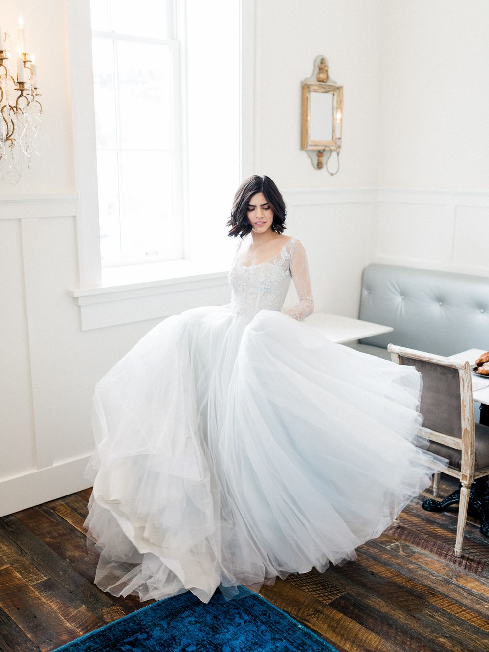 A bride in a flowing tulle gown twirls gracefully across a rustic wooden floor in a bright white room.