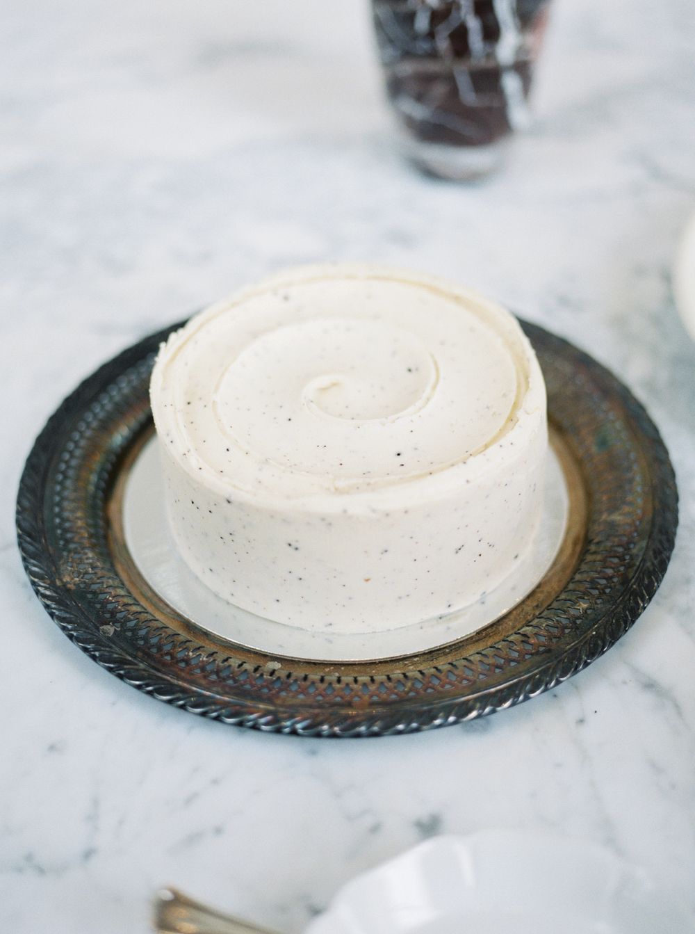Simple white frosted cake on decorative silver plate against marble surface.