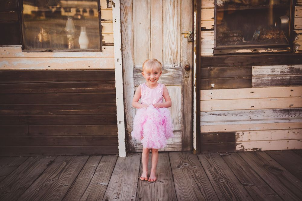 A small child in a pink tutu dress stands on a rustic wooden porch in front of a weathered door near Cherry Valley, Ca.