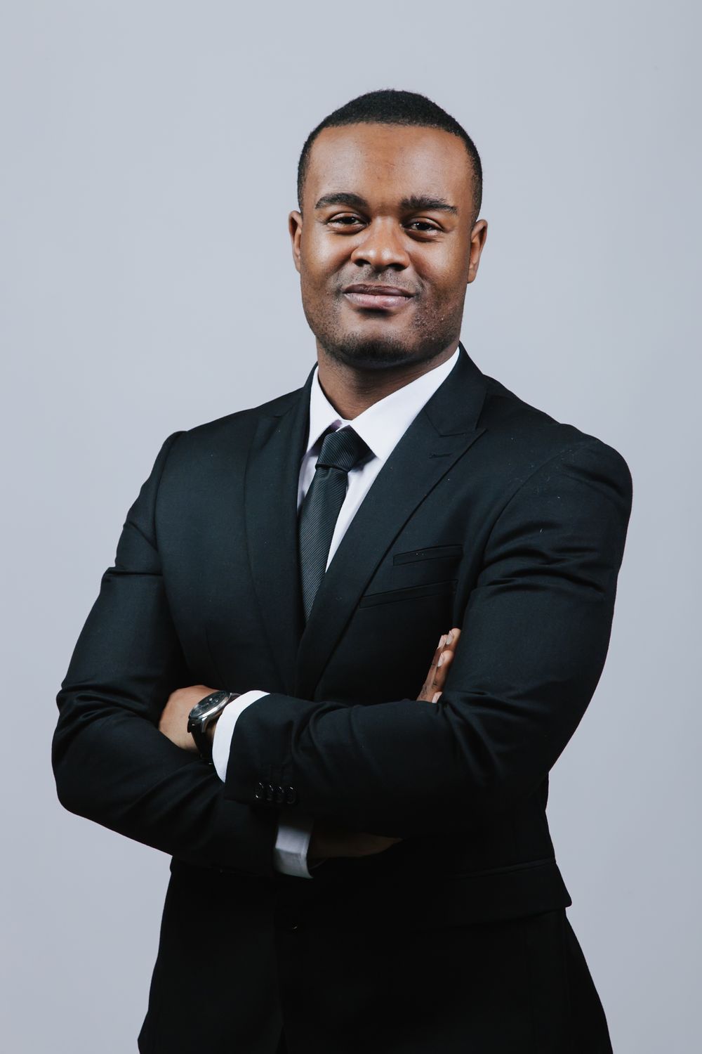 Professional headshots of someone in a black suit and tie posing with crossed arms against a light gray background.