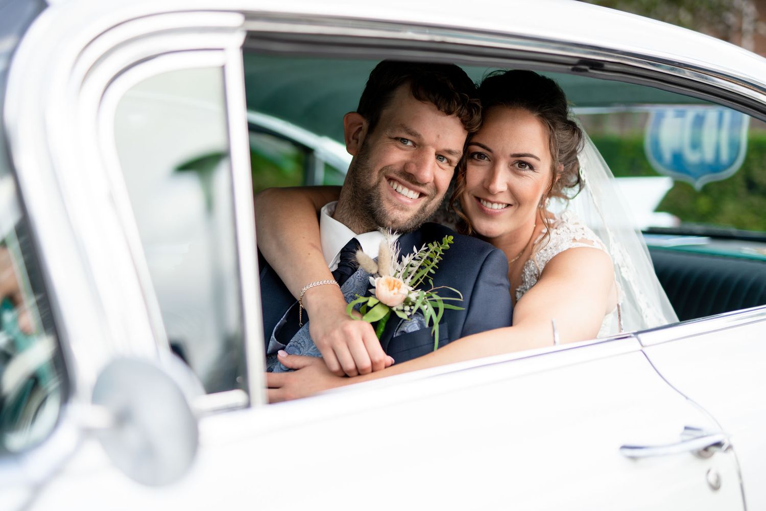 Happy newlyweds smile together while seated in a classic white car decorated with wedding flowers.
