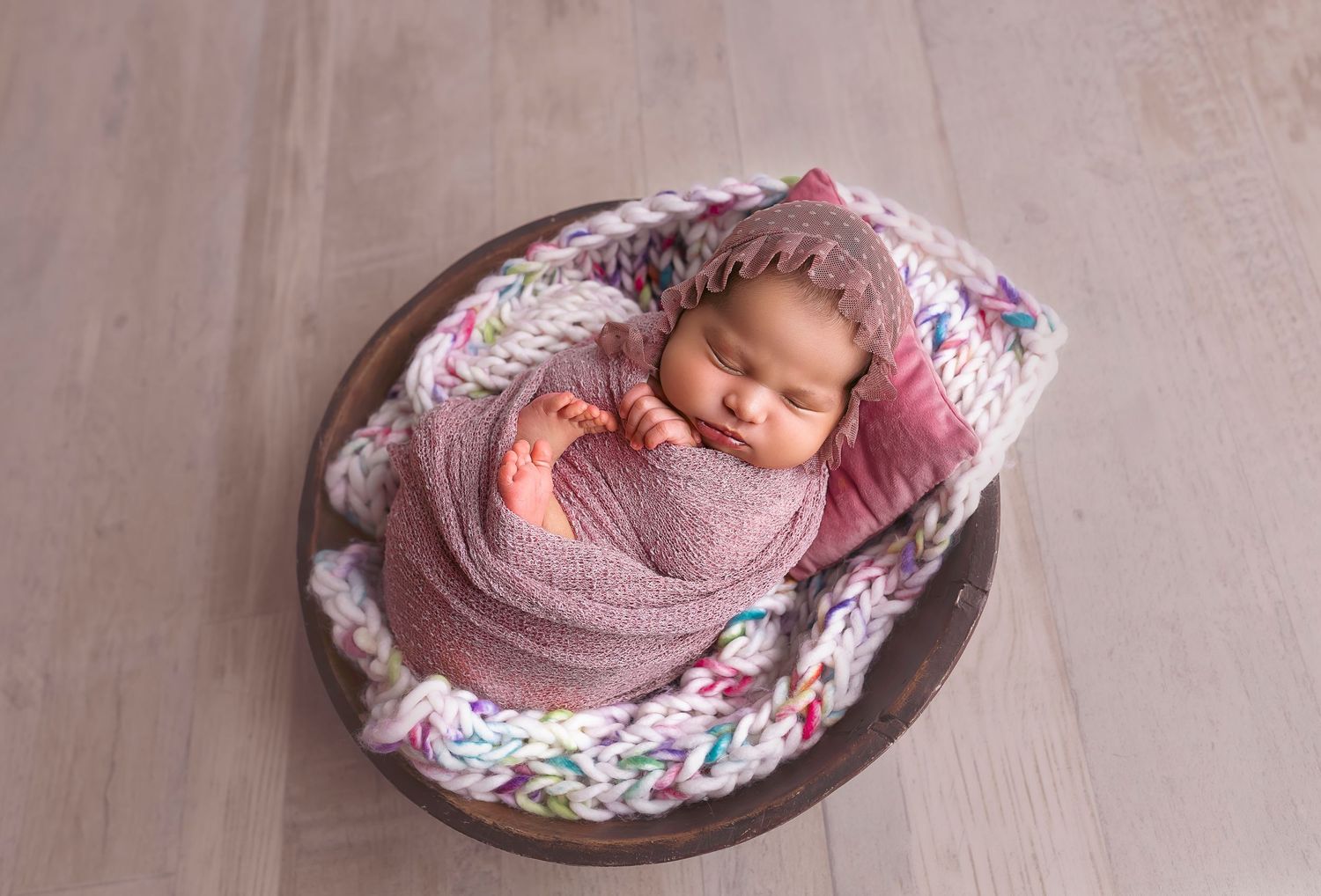 bebe recien nacida dentro de un bowl redondo color marron de madera y envuelta en una manta lavanda y gorrito purpura