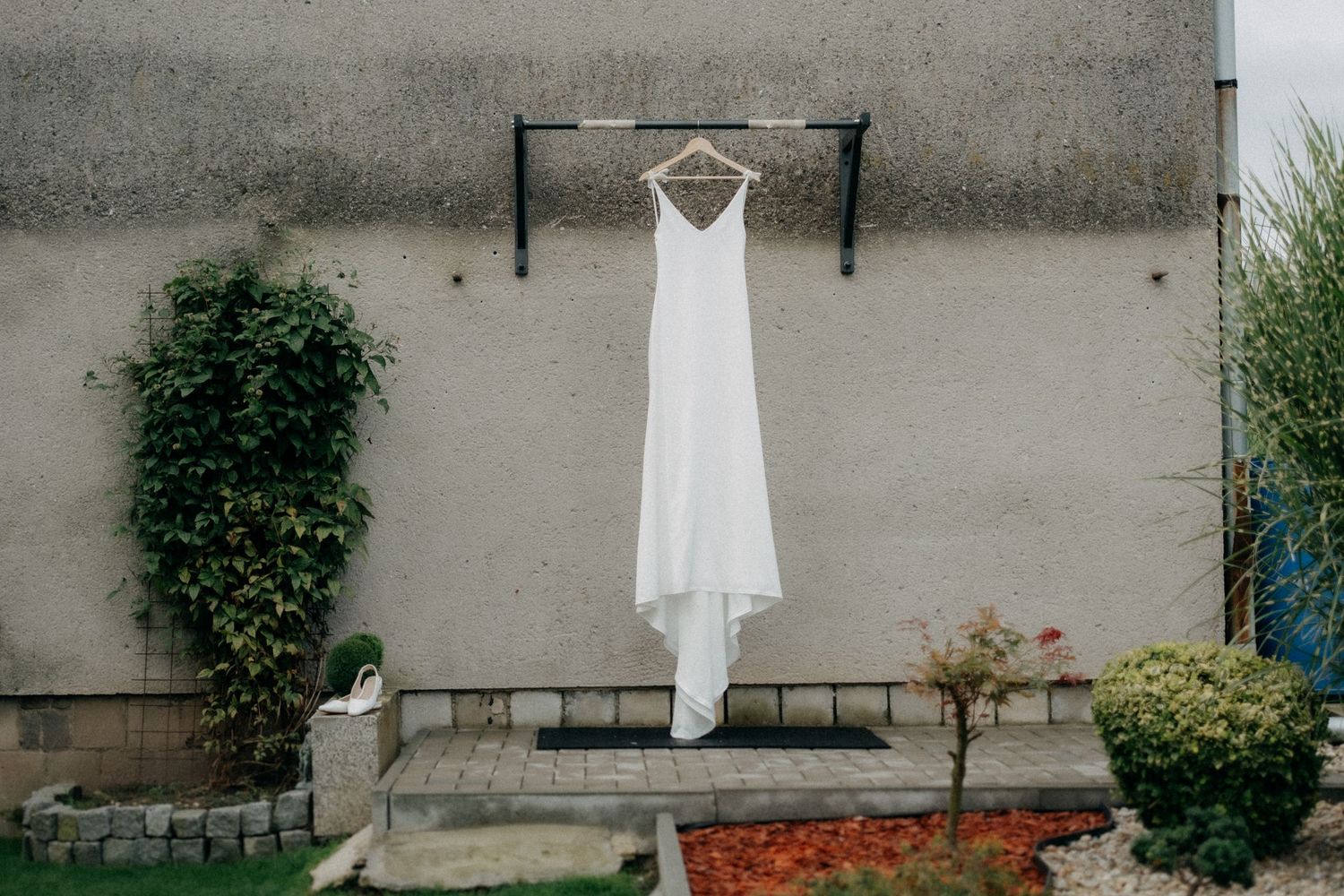 White wedding dress hangs elegantly against a gray concrete wall with climbing vines.