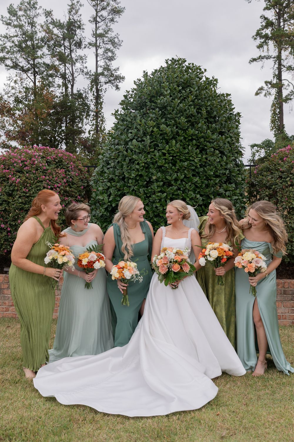 A wedding party wearing sage green and mint colored dresses poses together outdoors with colorful bouquets.