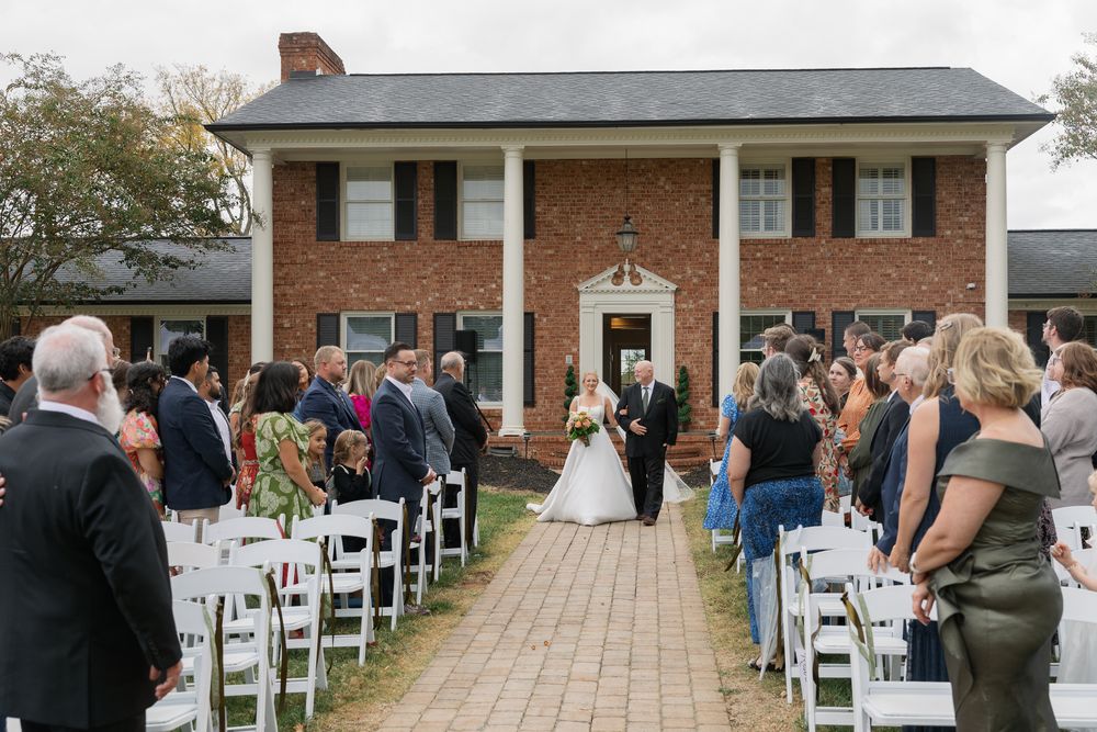 Outdoor wedding ceremony takes place on brick walkway in front of traditional colonial style brick house with white columns.