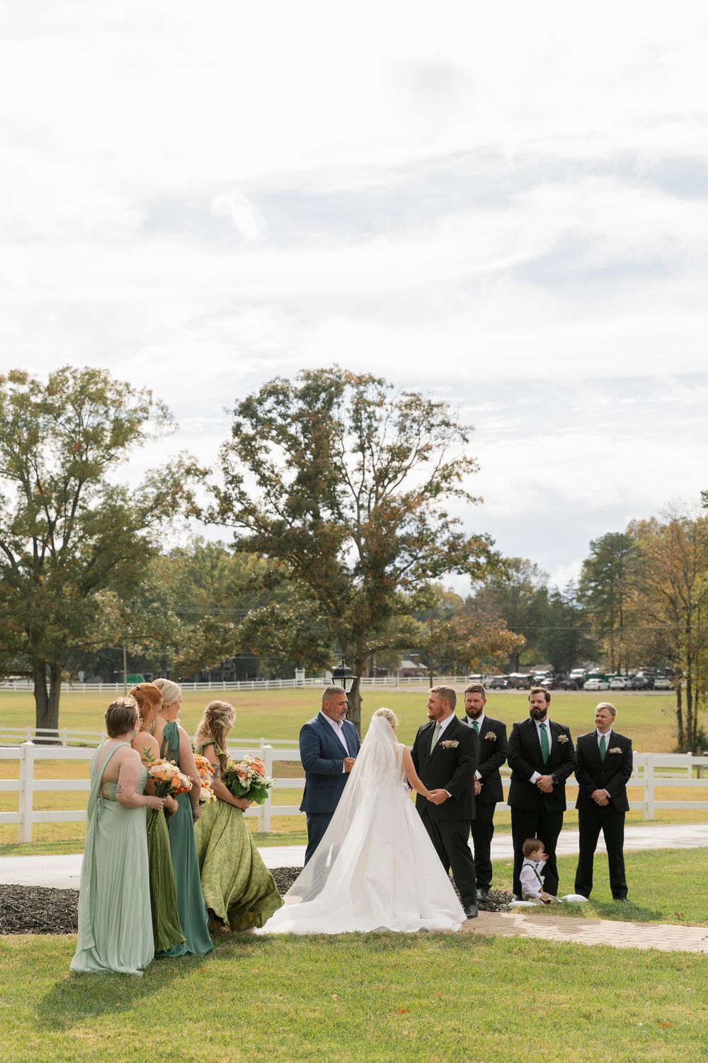 A wedding party stands outdoors on a grassy lawn with white fences and autumn trees in the background.