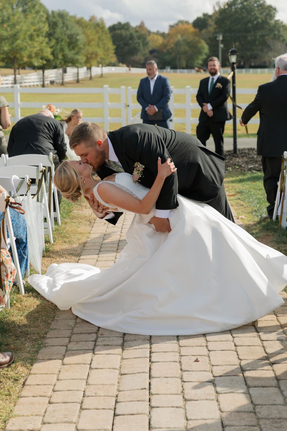 A couple shares a romantic dip kiss during their wedding ceremony on a stone pathway surrounded by white chairs.