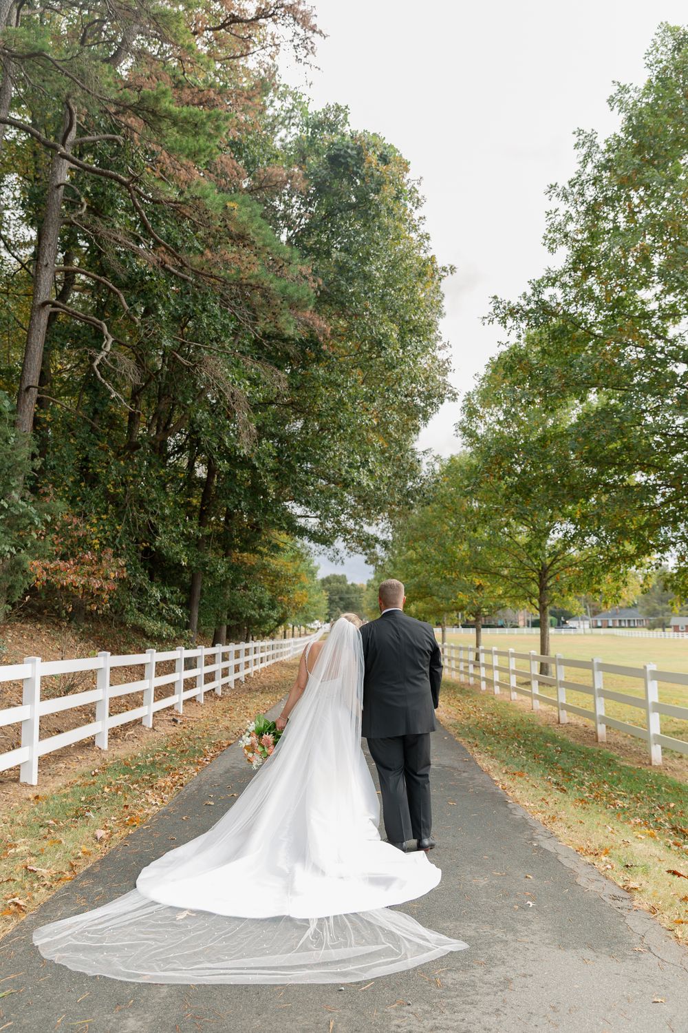 A bride and groom walk along a tree-lined path with white fencing on an autumn day.