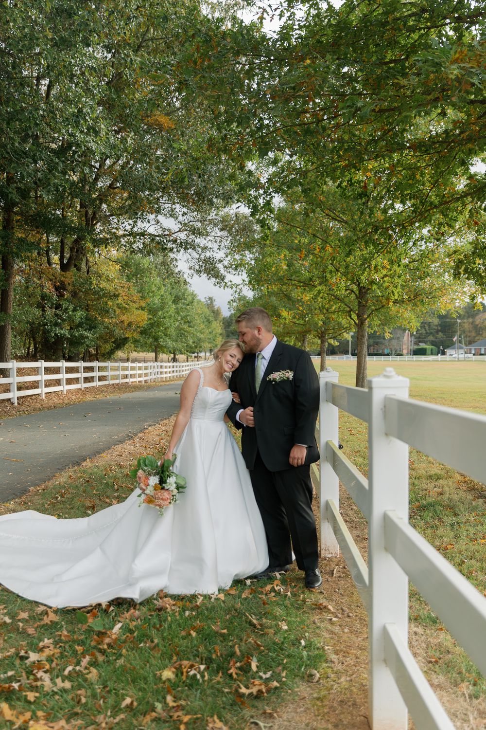 A romantic wedding photo along a white fence-lined path with fall foliage and trees in the background.