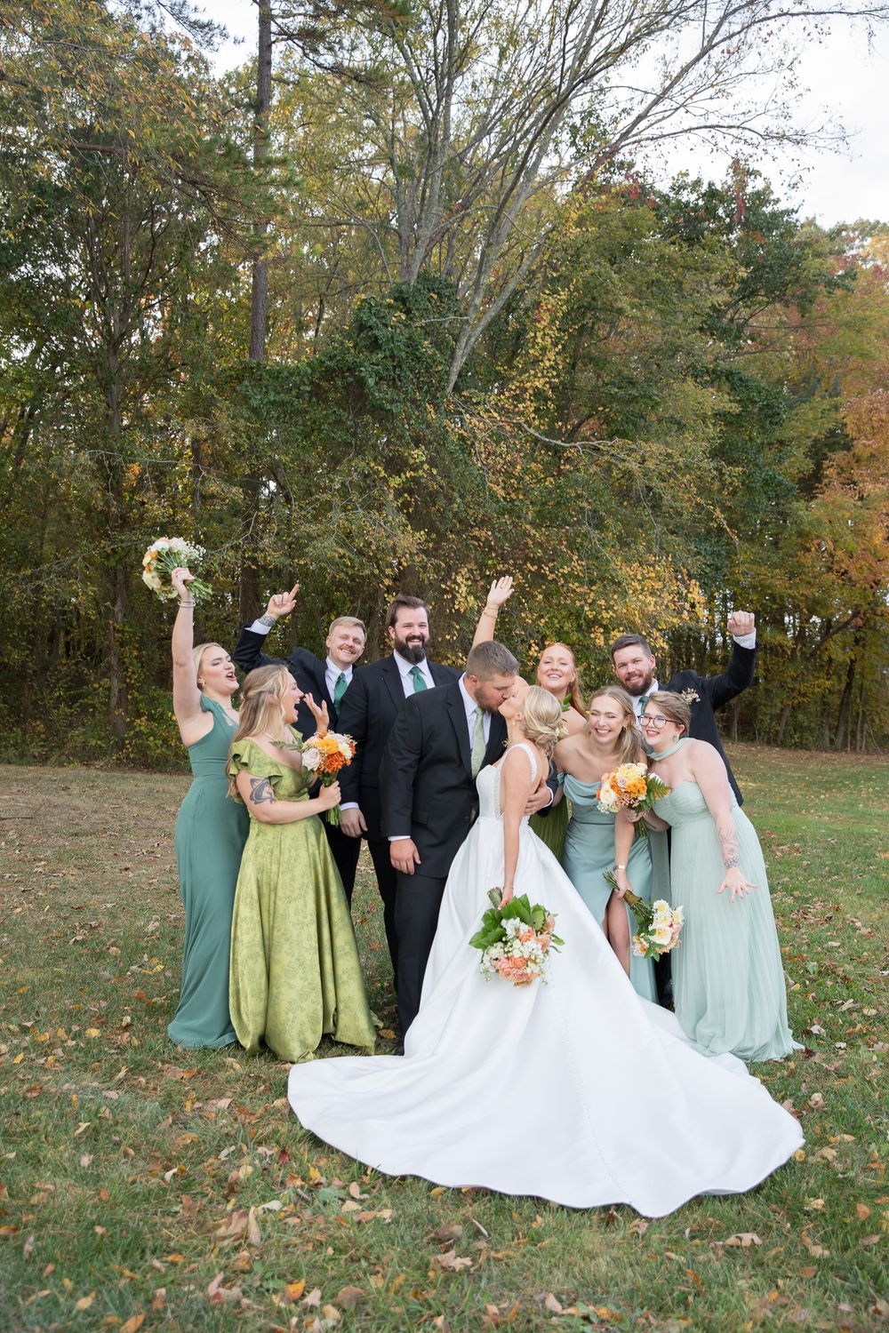 A wedding party poses joyfully on grass with autumn trees behind them in pastel bridesmaid dresses.