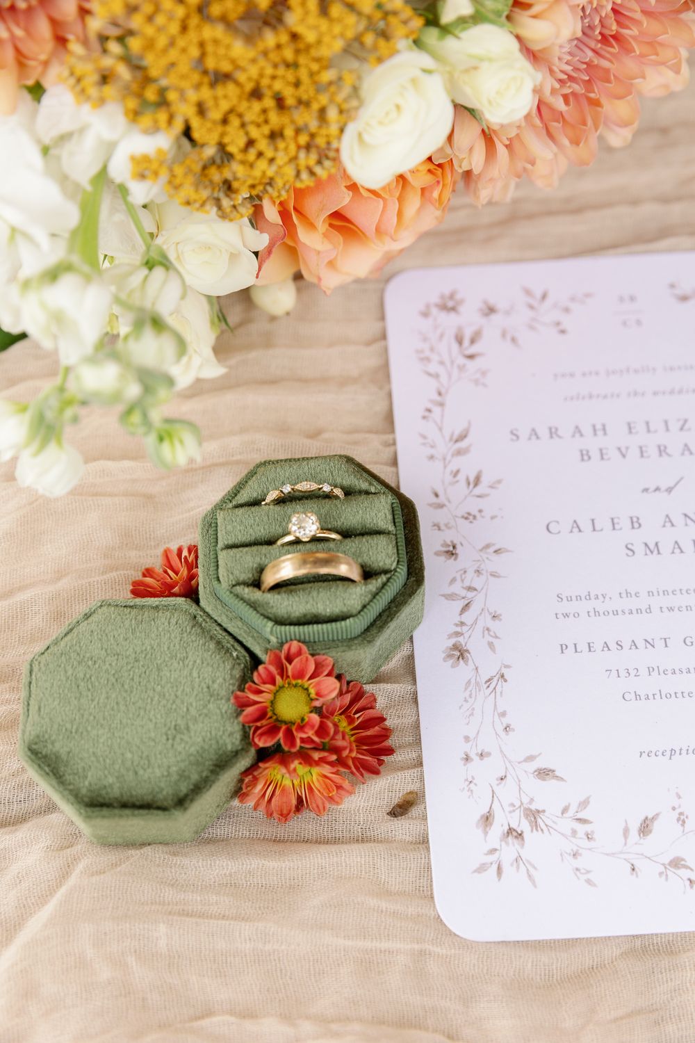 Wedding invitation and engagement ring in sage green velvet box with coral and white flowers on beige fabric.