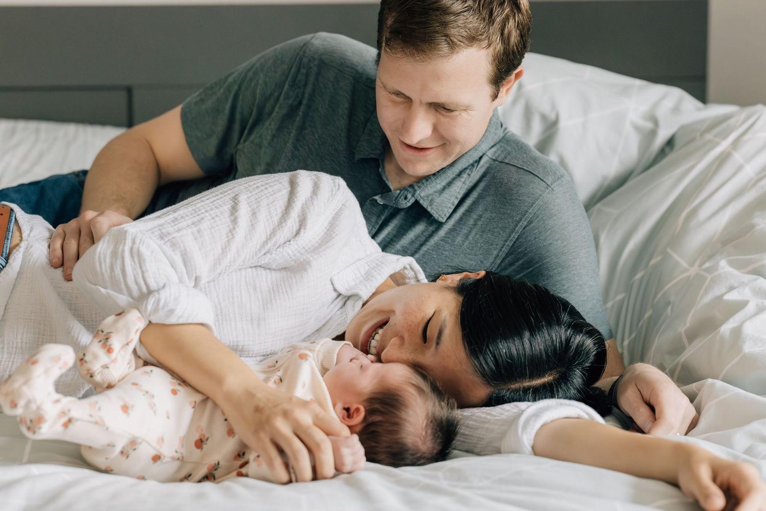 Couple laughing and cuddling with newborn baby on white bed during in-home newborn session.