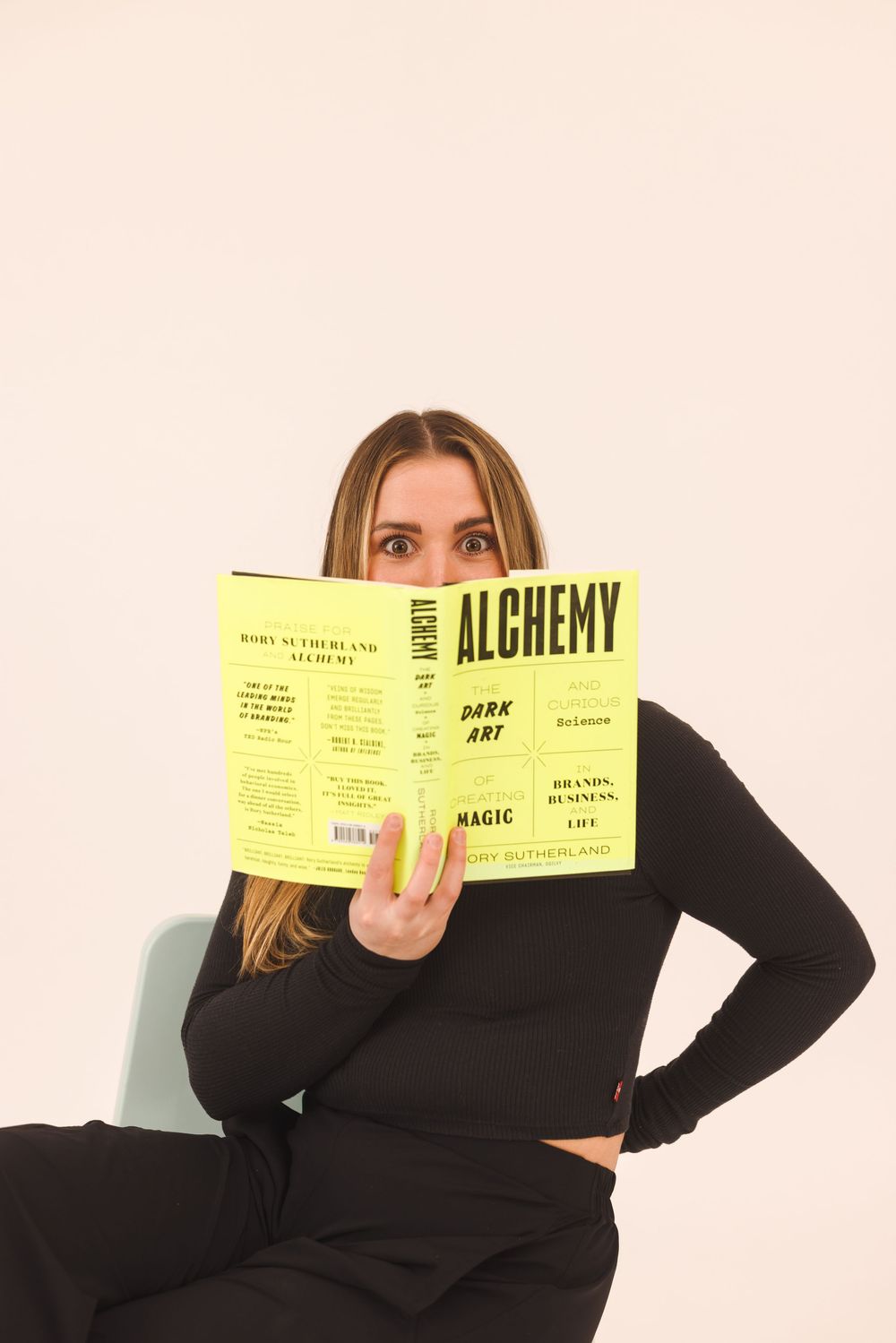 Person in black sweater holds and poses with bright yellow book titled Alchemy in different playful positions.