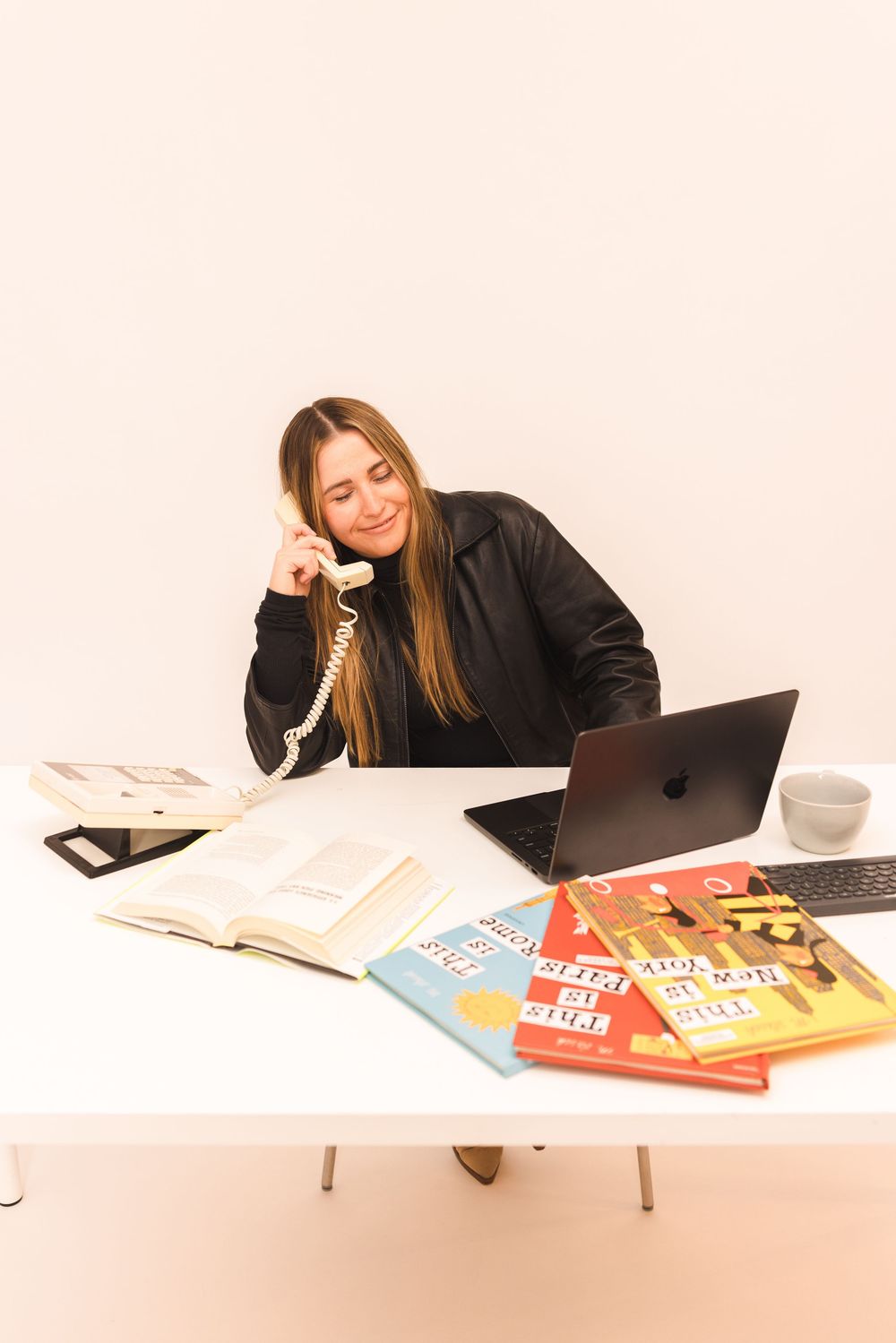 Student in black jacket studies at desk with laptop and colorful books spread out on white table.