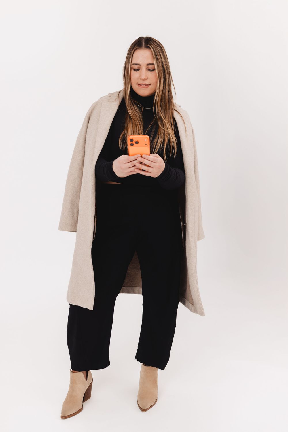 Stylish minimalist fashion portrait showing a black turtleneck, beige coat and boots against white background.