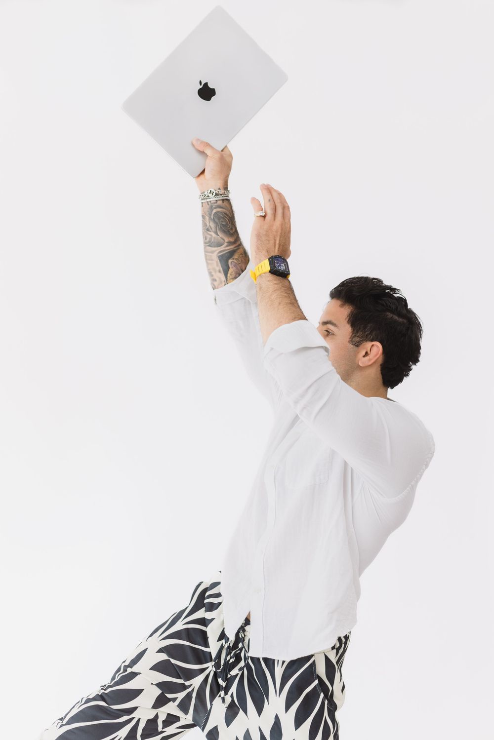 Person in white shirt with tattoos holding up a MacBook laptop against white background with black floral pattern pants.
