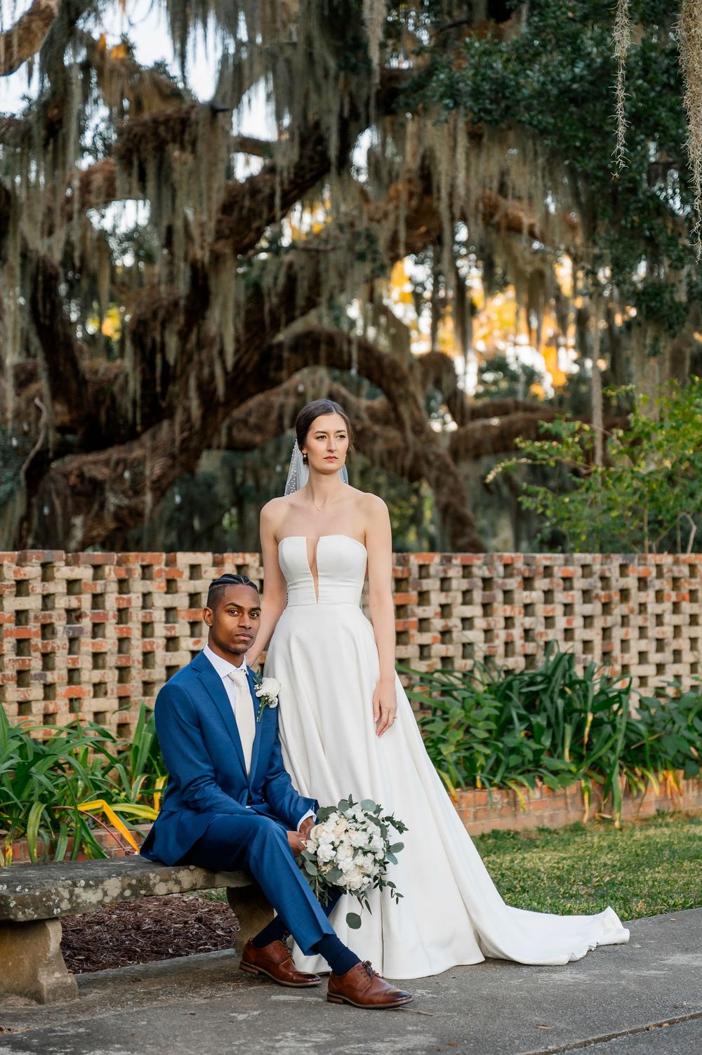A romantic wedding photoshoot in a garden with Spanish moss draped trees and a decorative brick wall.