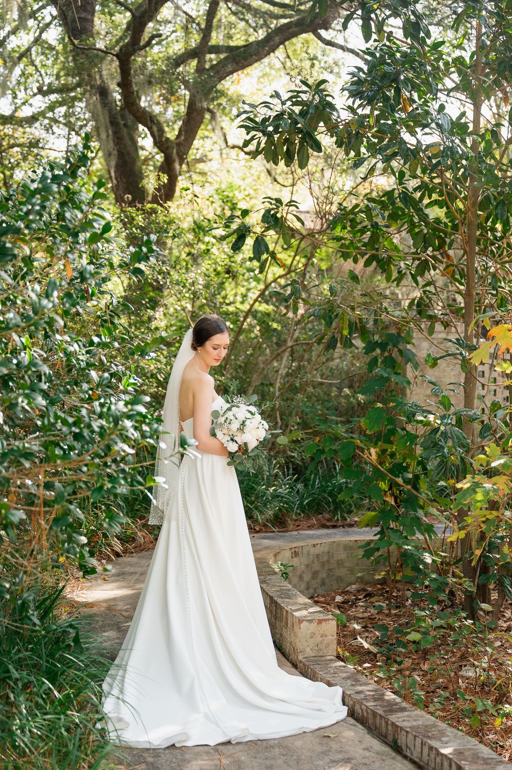 Bride in flowing white gown poses gracefully in a lush garden setting with brick pathways.
