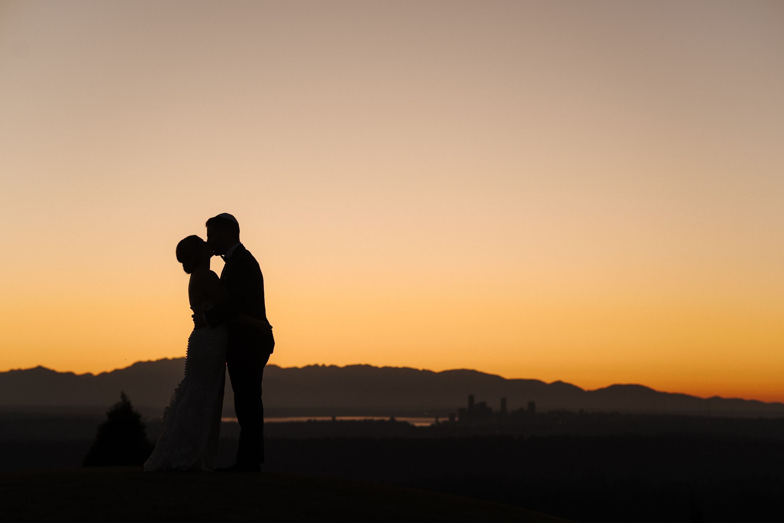 Silhouetted figures stand together against a vibrant orange and purple sunset sky with mountain ridges in the background.