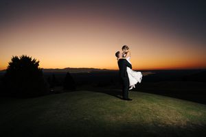 A silhouetted figure sits on a grassy hilltop against a dramatic sunset sky with orange and purple hues.