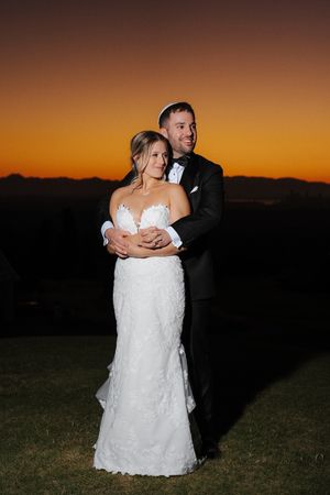 A bride and groom embrace on a grassy hill at sunset with a vibrant orange sky behind them.